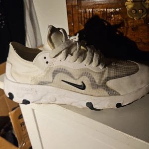 Nike React Element 55 vita sneakers - Nike React Element 55 sneakers i vitt med svarta detaljer och genomskinlig mesh på ovandelen. Snygg vågig design på sidorna, klassisk snörning och bekväm, dämpad sula med svarta inslag. Perfekta för dig som gillar sportig och modern stil.