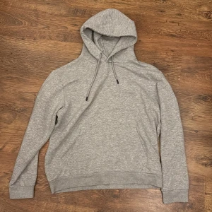 Grå hoodie från Jack & Jones - Klassisk grå hoodie från Jack & Jones med huva och snörning. Tröjan har en känguruficka framtill och ribbade muddar vid ärmslut och nederkant. Perfekt för chill dagar och enkel att matcha med jeans eller joggers.