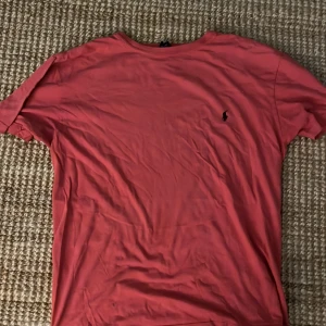 Korallröd t-shirt från Polo Ralph Lauren - Säljer en korallröd t-shirt från Polo Ralph Lauren med klassisk svart broderad logga på bröstet. T-shirten är i 100% bomull och har en enkel, stilren design med rund hals och korta ärmar. Perfekt för dig som gillar en clean och tidlös look.