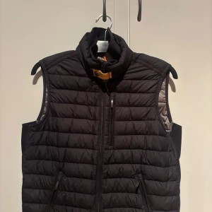 Svart dunväst från Parajumpers - Snygg svart dunväst från Parajumpers med quiltad design och hög krage med gul spänndetalj. Västen har dragkedja framtill och två fickor med dragkedja. Insidan är grå och har märkets logga. Perfekt för lager-på-lager under kyliga dagar.  Västen är köpt på NK i Göteborg, kvitto finns som kommer med vid köp! 