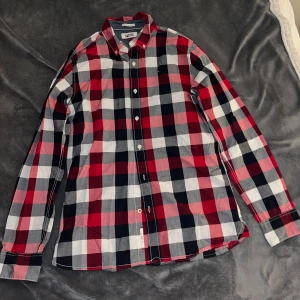 Rutig långärmad skjorta från Tommy Hilfiger - Snygg rutig skjorta från Tommy Hilfiger i rött, vitt, grått och mörkblått. Klassisk button-down krage och vita knappar framtill. Skjortan är tillverkad i mjuk bomull och har lång ärm med manschett. Perfekt för dig som gillar stilrena och färgglada plagg. Passar fint till julen som närmar sig runt hörnet. 