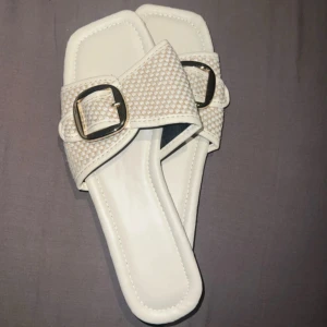 Vita sandaler storlek 38 helt nya - Snygga vita sandaler med fyrkantig tå och bred rem i rutigt mönster. Dekorativt metallspänne framtill ger en trendig touch. Sulan är platt och svart undertill, ovandelen är i syntetmaterial. Perfekta för varma dagar och enkel att matcha med sommarens outfits.