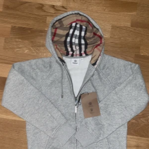 Grå hoodie från Burberry med dragkedja - Snygg grå hoodie från Burberry med klassiskt rutigt foder i huvan. Tröjan har dragkedja framtill, snörning i huvan och ribbade muddar. Perfekt för dig som vill ha en stilren och exklusiv look med en touch av Burberrys ikoniska design.