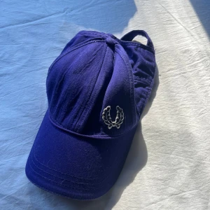 Lila keps från Fred Perry i bomull - Snygg lila keps från Fred Perry i 100% bomull. Klassisk modell med böjd skärm, justerbar rem baktill med metallspänne och broderad Fred Perry-logga framtill. Perfekt accessoar för att lyfta din stil.