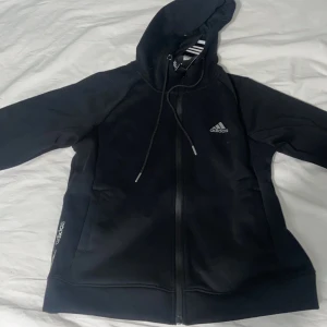 Svart hoodie från Adidas med dragkedja - Svart hoodie från Adidas med hel dragkedja framtill och justerbar huva med snörning. Klassisk Adidas-logga på bröstet och diskret logga vid fickan. Perfekt för en sportig och avslappnad stil.
