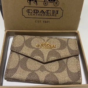 Coach plånbok i beige och brunt - Snygg plånbok från Coach med klassiskt monogrammönster i beige och brunt. Plånboken har guldfärgad logga framtill, kuvertformad flik och flera fack på insidan. Tillverkad i tåligt skinnmaterial och levereras i originalask.
