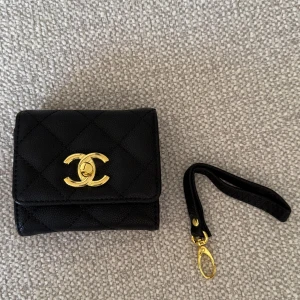 Plånbok  - Snygg svart plånbok i skinn med guldigt spänne framtill. Kommer med avtagbar handledsrem i matchande svart skinn och gulddetaljer. Flera kortfack och plats för mynt och sedlar. Perfekt accessoar för dig som gillar lyxiga detaljer.