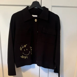 Svart overshirt från Sandro med tryck - Svart overshirt från Sandro med coola gula tryck och broderier. På ryggen finns en stor rund patch med smiley och texten 'Give Back Love', samt samma text broderad på ena ärmen. Jackan har två bröstfickor, knappar framtill och klassisk krage.