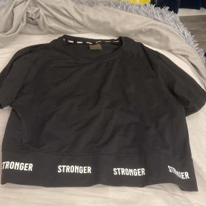 Svart croppad t-shirt från Stronger - Svart croppad t-shirt från Stronger med vit logga tryckt flera gånger längs nederkanten. Tillverkad i stretchigt material med 90% återvunnen polyester och 10% elastan. Perfekt för träning eller chill dagar. Kortärmad och med rund hals.