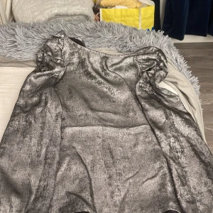 Glittrig silverklänning från Lindex - Snygg kort glittrig klänning från Lindex i silver med puffiga axlar och lång ärm. Perfekt för dig som vill sticka ut med metallic-look och coola detaljer. Klänningen har en rak passform och är tillverkad i ett glansigt material.