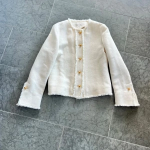 Vit kavaj från By Malene Birger - Stilren vit kavaj från By Malene Birger med fransade kanter och guldknappar framtill och vid ärmslut. Jackan har en rak passform, rund halsringning och är tillverkad i ett strukturerat tyg som ger en exklusiv känsla. Oanvänd men tyvärr ingen prislapp kvar. Nypris 8000kr. Vid snabb affär kan jag gå ner till 800.