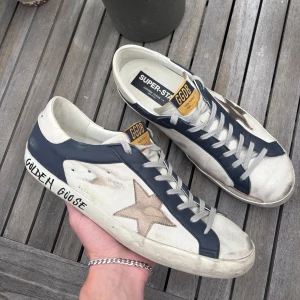 Golden Goose Superstar sneakers blå/vit - Golden Goose Superstar sneakers i vitt och mörkblått skinn med beige stjärndetalj på sidan. Klassisk låg modell med snörning, svart sula och handskriven logga på sidan. Tungan har gul etikett med GGDB-tryck. Perfekta för en avslappnad streetstil. Skorna är köpta nya på NK i Stockholm för 5399kr. Kvitto finns. Priset är inte hugget i sten!