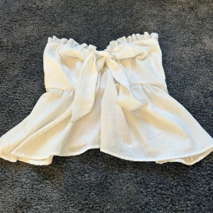 Vit axelbandslös tubtopp med knyt - Söt vit tubtopp med volangkant upptill och knyt framtill. Toppen är axelbandslös och har en lätt struktur i tyget samt smock baktill för extra komfort. Perfekt för en somrig look och passar till både jeans och kjol.