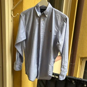 Ljusblå skjorta från Ralph Lauren - Klassisk ljusblå skjorta från Ralph Lauren i 100% bomull. Skjortan har button-down krage, långa ärmar och den ikoniska broderade loggan på bröstet. Perfekt för en clean och stilren look. Använd 1 gång.