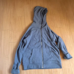 Grå hoodie med dragkedja och huva (Alldrig Använd) - En basic grå hoodie från 157 med hel dragkedja framtill och stor huva. Tröjan har känguruficka och ribbade muddar vid ärmslut och nederkant. Perfekt för chill dagar eller när du vill ha något bekvämt och enkelt.