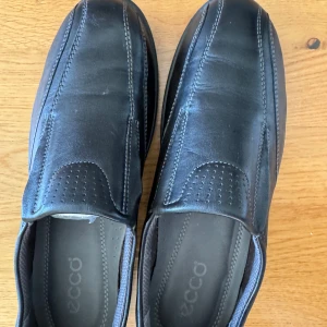 Svarta loafers från Ecco i skinn - Stilrena svarta loafers från Ecco i mjukt skinn med diskreta sömmar och perforerade detaljer framtill. Skorna har en platt sula och rund tå, perfekt för en clean och klassisk look. Ecco-logga bak på hälen. Snygga och bekväma för vardagsstilen.