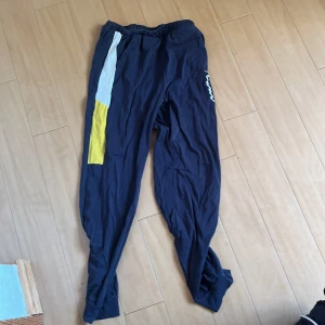 Blå SHEIN mjukisbyxor med färgblock - Säljer ett par blå mjukisbyxor från SHEIN med coola färgblock i vitt och gult på ena benet. Byxorna har elastisk midja och en avslappnad passform, perfekta för chill dagar eller träning. Materialet är mjukt och skönt mot huden.