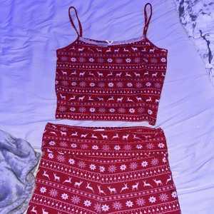 Röd julig linnetopp med spets - Supersöt röd linnetopp med smala axelband och vitt julmönster med renar, snöflingor och granar. Toppen har vit spetskant och en liten rosett framtill. Perfekt för mysiga vinterkvällar eller julfirande.