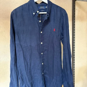 Marinblå linneskjorta Polo Ralph Lauren - Snygg marinblå skjorta från Polo Ralph Lauren i linne med klassisk krage, vita knappar och röd broderad logga på bröstet. Custom fit-modell med lång ärm, perfekt för en stilren och avslappnad look.