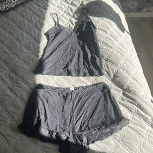 Svart linnetopp med spetsdetaljer - Svart linnetopp med tunna axelband och v-ringning, dekorerad med spets längs urringningen. Tillverkad i mjukt och lätt material, perfekt för en chill look. Matchande shorts med spetskant ingår.