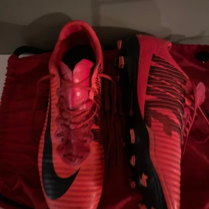 Nike Mercurial elits  fotbollsskor - Säljer ett par Nike Mercurial fotbollsskor i rött med svarta detaljer och snörning. Skorna har en smal, sportig form och mönstrade svarta linjer på sidorna. Yttersulan är svart med röda dobbar, perfekt för dig som vill sticka ut på planen.