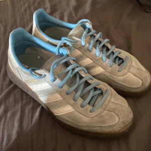 Adidas Originals Schweiz sneakers 38 - Säljer ett par Adidas Originals Schweiz sneakers i ljusgrå mocka med vita ränder och ljusblå detaljer. Skorna har klassisk gummisula, snörning och Adidas-logga i guld på sidan. Perfekt för dig som gillar retrostil och snygga färgkombos.