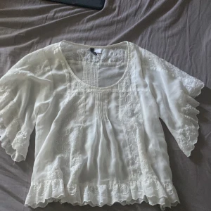 Vit broderad blus från H&M Divided - Söt vit blus från H&M Divided med broderade detaljer och volangkant. Blusen har korta, vida ärmar och rund halsringning. Perfekt för en romantisk look med sina spets- och broderimönster. Luftigt material som passar bra till jeans eller kjol.