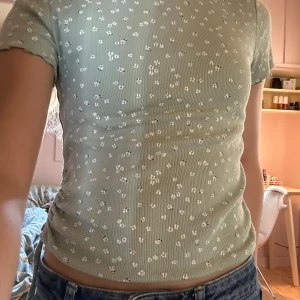 Grön blommig ribbad topp från H&M - Söt ljusgrön ribbad topp från H&M med små vita blommor och korta ärmar. Toppen har rund halsringning och snörningsdetalj i sidan som ger en snygg rynkad effekt. Tröjan är lite trådig på vissa ställen men det är inget som man tänker på! Kan tänka mig att sänka priset om någon är intresserad!!