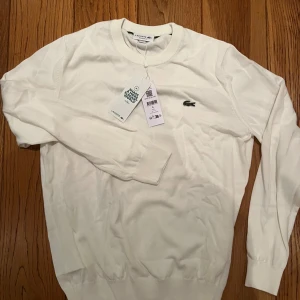 Vit tröja från Lacoste, regular fit - Vit långärmad tröja från Lacoste med klassisk rund halsringning och den ikoniska krokodilloggan broderad på bröstet. Tröjan är i regular fit och tillverkad av bomull och polyamid för en mjuk och skön känsla. Aldrig använd 