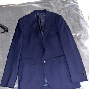 Mörkblå kostym från Riley i ull - Säljer en stilren mörkblå kostym från Riley i ull. Kavajen har två knappar, klassiska slag och innerfoder i blankt tyg. Byxorna är raka med pressveck och hällor för bälte. Perfekt för dig som vill ha en clean och tidlös look. Har enbart amvänds vid ETT tillfälle och därefter gått igenom kemtvätt. Köpta från Brothers. Nypriset i butik för hela kostymen: 3000kr. 