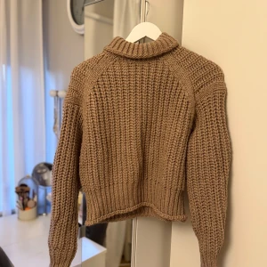 Beige stickad polotröja - Säljer en beige stickad polotröja med grov ribbstickning. Tröjan har lång ärm och en hög krage som ger en mysig och trendig look. Perfekt att styla med jeans eller kjol för en fin vardagsstil.