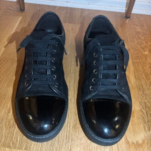 Svarta sneakers från Lanvin i mocka - Snygga svarta sneakers från Lanvin med ovandel i mocka och lackad tå. Skorna har svarta snören, metallöljetter och en platt sula. Klassisk och stilren design som passar till det mesta. Perfekta för dig som gillar exklusiva och tidlösa sneakers. Skorna är hela och  i bra skick. Vanligt bruksslitage. Skriv om du undrar något eller vill ha fler bilder!