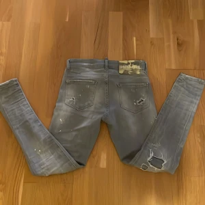 Grå  jeans från Dsquared2 - Säljer ett par grå  jeans från Dsquared2 med slitningar, hål och färgstänk för en riktigt edgy look. Jeansen har klassisk femficksdesign, låg midja och Dsquared2-logga på bakfickan och vid gylfen. Perfekta för dig som gillar streetstyle och detaljer.