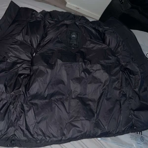 Svart pufferväst från Canada Goose - Säljer en svart pufferjacka från Canada Goose i storlek M. Jackan har klassisk quiltad design, dragkedja framtill och ikonisk logga på bröstet. Perfekt för kalla vinterdagar och riktigt snygg till streetwear-stilen.