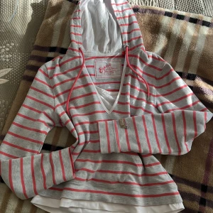 Randig hoodie  - Snygg hoodie från Esprit i grått och vitt med röda ränder. Tröjan har huva med snörning, v-ringad hals och långa ärmar. Mjuk bomullskvalitet och coolt tryck på bröstet. Perfekt för chill dagar och enkel att matcha med jeans eller leggings.