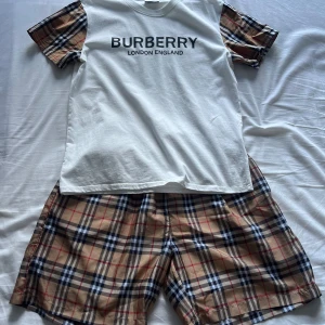 Burberry t-shirt och shorts (suit) - Snygg vit t-shirt från Burberry med klassiska rutiga ärmar i beige, svart och rött. Stor Burberry-logga framtill. T-shirten har korta ärmar och är gjord i mjuk bomull. Perfekt för dig som vill sticka ut med en ikonisk look.