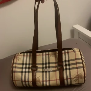 Rutig handväska från Burberry - Säljer en klassisk handväska från Burberry i beige med brunt, svart och rött rutmönster. Väskan har bruna skinnhandtag och detaljer, cylinderformad design och dragkedja upptill. Perfekt storlek för att få plats med allt du behöver. Inte använd alls! 