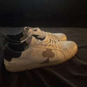 Vita sneakers från The Gallery Club - Snygga vita sneakers i skinn från The Gallery Club med svart häl och grått klövermotiv på sidan. Klassisk rund tå, vita snören och platt sula. Perfekta för dig som gillar stilrena detaljer med en twist.