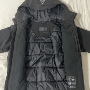 Svart dunjacka från Sail Racing - Svart dunjacka från Sail Racing med huva och två bröstfickor med dragkedja. Jackan har quiltad insida, hög krage och logga på bröstet. Perfekt för kalla dagar och har en clean, sportig look.