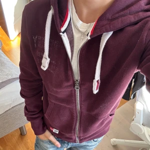 Vinröd hoodie - Säljer en vinröd hoodie  med dragkedja, vita snören och huva. Tröjan har två fickor framtill och diskret broderad logga på bröstet. Perfekt för chill dagar och snygg till jeans. Står att det är storlek S men den är större i storleken så M tycker jag då denna är stor för mig. Priset är ej hugget i sten.