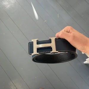 H bälte - Snyggt svart skärp från Hmed ett stort, silverfärgat H-spänne. Bältet är gjort i skinn med vit kontrastsöm längs kanterna som ger en clean och exklusiv vibe. Perfekt accessoar för att lyfta din outfit med en lyxig touch.