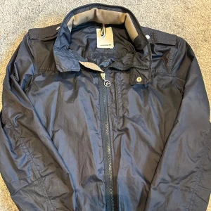 Mörkblå vindjacka från J.Lindeberg - Mörkblå vindjacka från J.Lindeberg med dragkedja framtill, hög krage och inbyggd huva som går att gömma. Jackan har snygga axeldetaljer och ribbad insida på kragen. Perfekt för blåsiga dagar och stilren look.