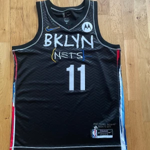 Brooklyn Nets Irving #11 Nike tröja - Säljer en svart baskettröja från Nike, Brooklyn Nets, med nummer 11 och Irving på ryggen. Designen är inspirerad av Jean-Michel Basquiat med färgglada detaljer längs sidorna och broderade loggor. Tillverkad i Dri-FIT-material för bästa komfort på planen samt streetwear.