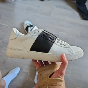 Valentino Open vita sneakers - Snygga vita Valentino Open sneakers med bred svart läderdetalj över snörningen. Klassisk rund tå och platt sula, diskret Valentino-logga på plösen och ikoniska nitar baktill. Perfekta för dig som gillar stilrena och exklusiva sneakers.