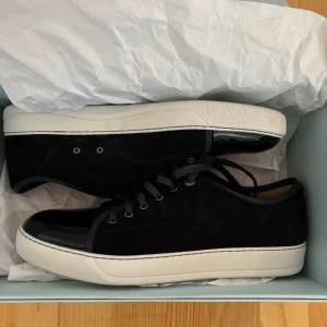 Svarta sneakers från Lanvin med lacktå - Snygga svarta sneakers från Lanvin med ovandel i mocka och en glansig lackad tå. Skorna har vita, tjocka gummisulor och svarta snören. Klassisk låg modell med rund tå och diskreta sömmar för en stilren look.