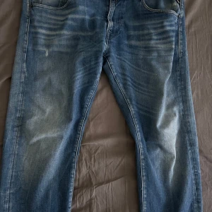 Blå Replay jeans i rak modell - Säljer ett par blå jeans från Replay i storlek 36. Jeansen har klassisk femficksdesign, snygga slitningar och ljusare partier framtill och baktill. Rak passform och normal midja. Perfekta för dig som gillar en avslappnad men stilren look.