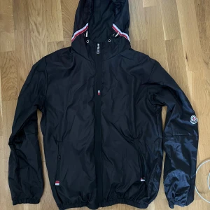 Svart vindjacka från Moncler - Svart vindjacka från Moncler med huva och dragkedja framtill. Jackan har Moncler-logga på ärmen och detaljer i rött, vitt och blått på huvan och vid fickorna. Tillverkad i lätt polyester, perfekt för blåsiga dagar.