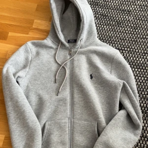 Grå hoodie från Polo Ralph Lauren - Snygg grå hoodie från Polo Ralph Lauren med dragkedja, huva och snörning. Klassisk broderad logga på bröstet och två fickor framtill. Perfekt för chill dagar och enkel att matcha med jeans eller joggers.