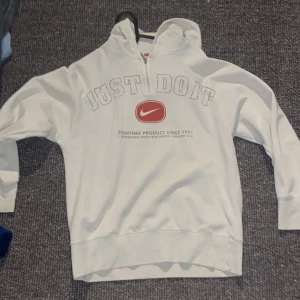 Vit Nike hoodie med tryck - Säljer en vit hoodie från Nike med halv dragkedja och huva. Framsidan har stort tryck med texten 'JUST DO IT' och röd Nike-logga. Perfekt för en sportig och avslappnad stil. Långärmad och mjuk i materialet.