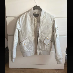 Vit bomberjacka från d.brand - Snygg vit bomberjacka från d.brand med guldiga dragkedjor och detaljer. Jackan har ribbade muddar vid ärmslut och midja, samt flera fickor med dragkedja och tryckknappar. Perfekt för dig som vill ha en clean och stilren look.